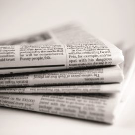 A close up black and white image of a stack four broadsheet newspapers with English text, casually piled up on a plain white background. The macro image is shot with a shallow depth of field, with only generic text visible.