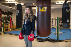 woman in business attire standing by a punching bag with red boxing gloves on