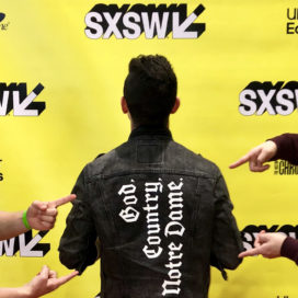 fingers pointing at the back of a jacket with the words God, Country, Notre Dame on it, in front of a yellow SXSW background