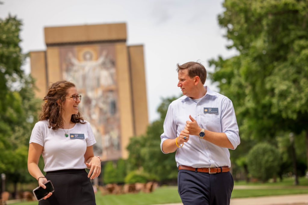 a man and woman walking with the hesburgh library in the background