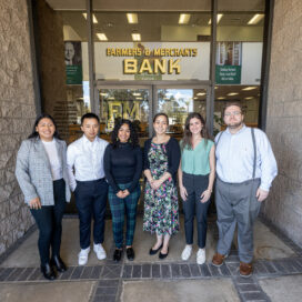 6 MNA students posing in front of a sign that reads "Farmers & Merchants Bank"