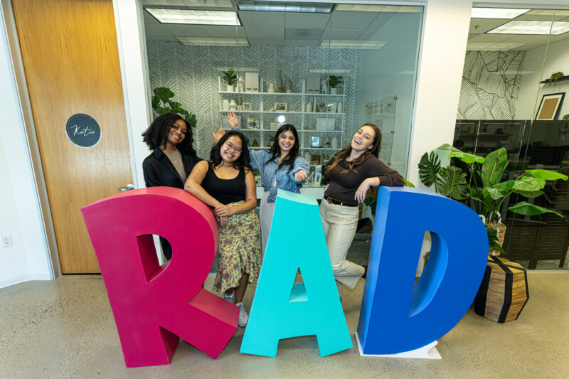 4 students in professional clothing posing behind a sign that reads "RAD"