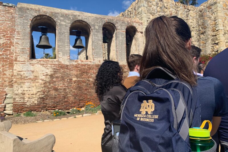 student with a Notre Dame Mendoza backpack standing in front of mission bells