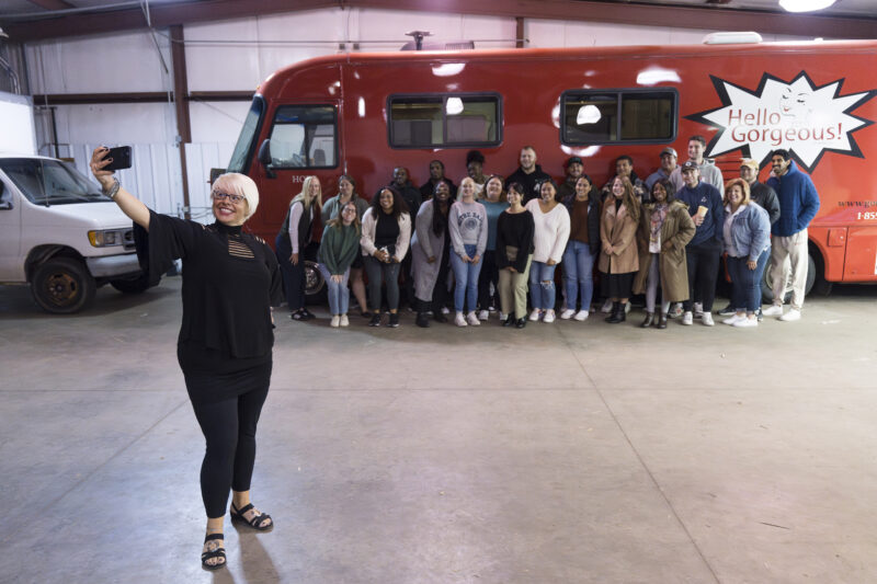 a woman with short platinum blond hair snaps a selfie of herself with a large group of students standing in front of a large red bus with a sign that reads "Hello Gorgeous!"