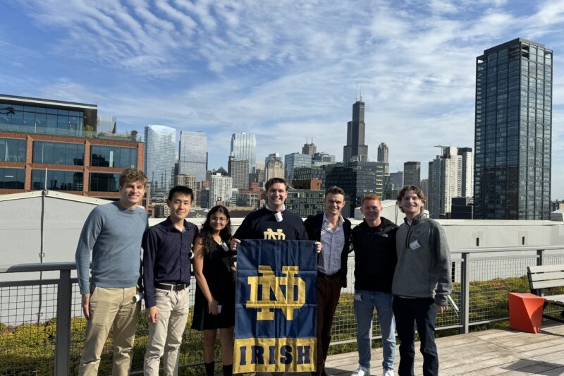 group of seven standing outdoors on a sunny day on a bridge overlooking a cityscape holding a blue and gold ND Irish flag