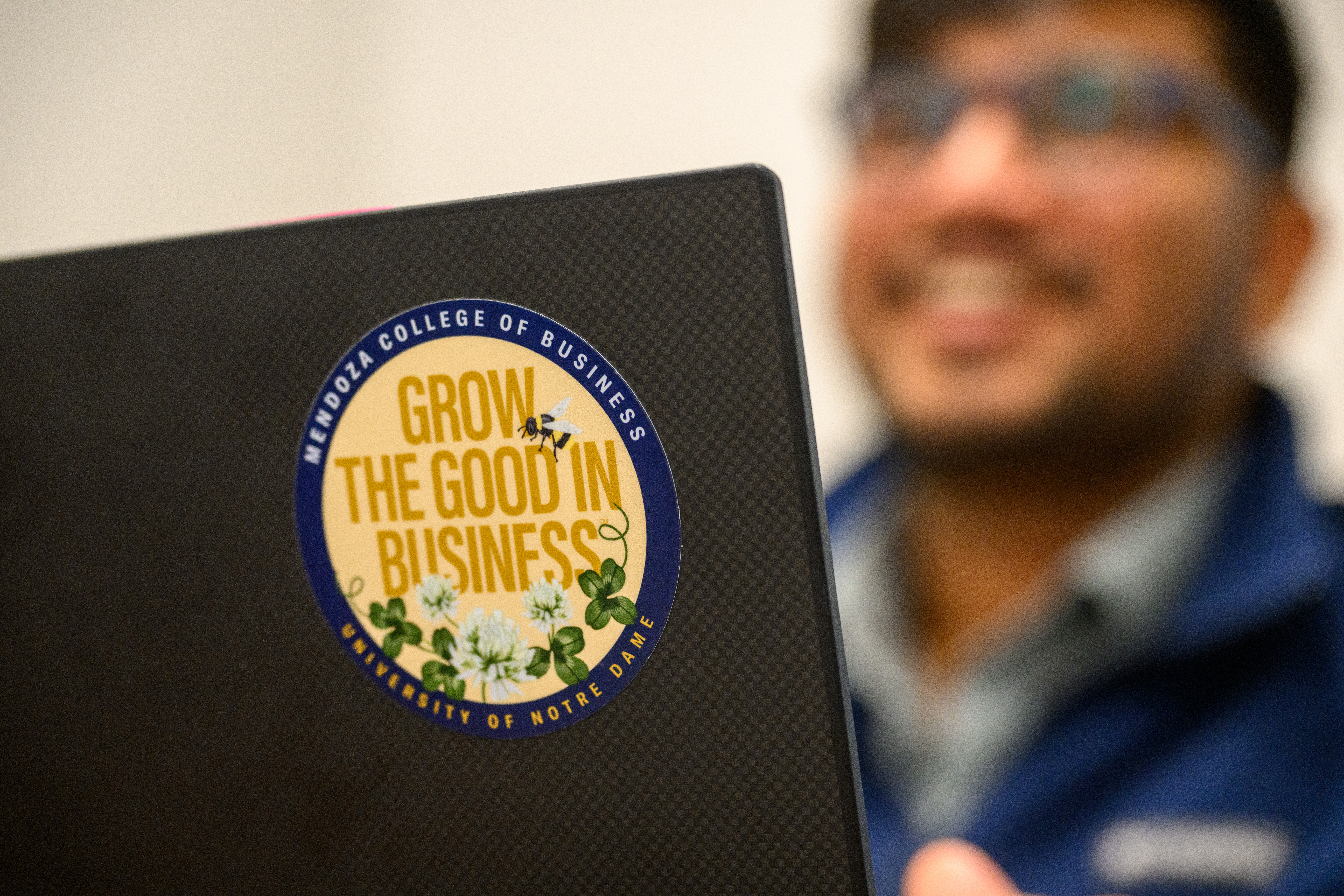 A student sits at a desk with a laptop in the foreground with a sticker reading "Mendoza College of Business: Grow the Good in Business"