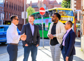 Four individuals stand outside in conversation on a sunny day, with urban surroundings in Dublin, Ireland including buildings and a colorful bus in the background. They are dressed in business casual attire, and one man is gesturing as he speaks while the others listen attentively.