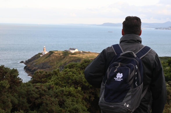 A man with a Mendoza College of Business backpack is standing in front of the cliffs in Ireland looking out at the water.