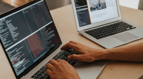 A student working on two laptops one with programming code and the other displaying an editor view of a website.