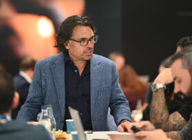 A man with shoulder-length hair, glasses, a blue blazer, and a dark shirt leans forward, engaged in conversation at a table with others. Beverages and papers are on the table, and the background is softly blurred.