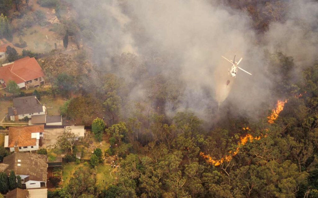 a helicopter flies over a wildfire in a forest next to homes.