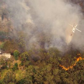 a helicopter flies over a wildfire in a forest next to homes.