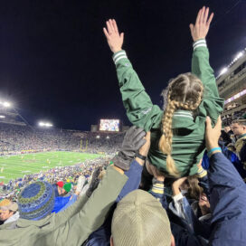 A group of students lifting up a female student who is cheering on the Notre Dame football team. Her arms are raised in the air as the lookout towards the field.