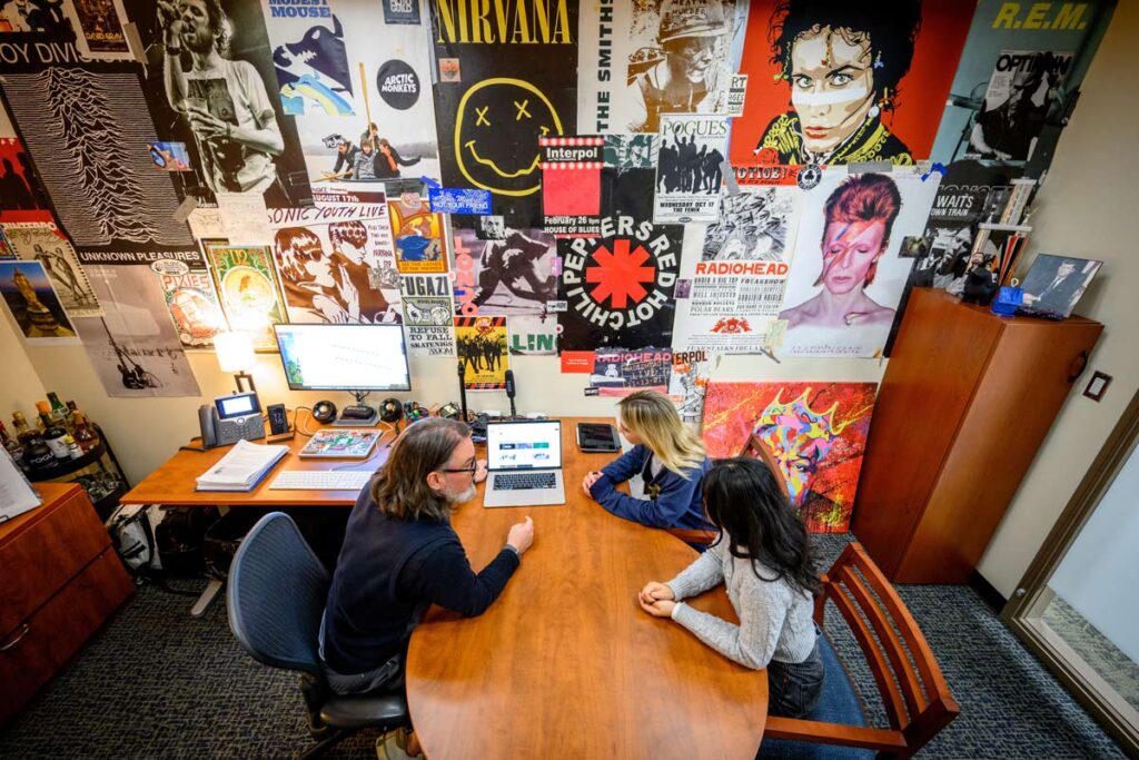 Kevin Hartman's office with posters all over the wall, looking down on Kevin with two students at his desk.