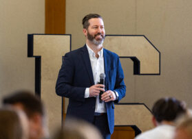 Notre Dame Professor Kris Muir teaching a class wearing a navy blue suit with the letters "IRISH" behind him.