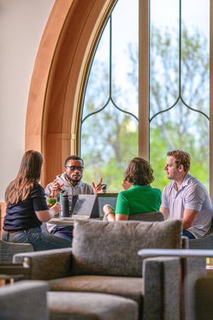 A group of four MBA students sit in front of a window in Stayer Center and discuss classwork with their laptops in front of them.