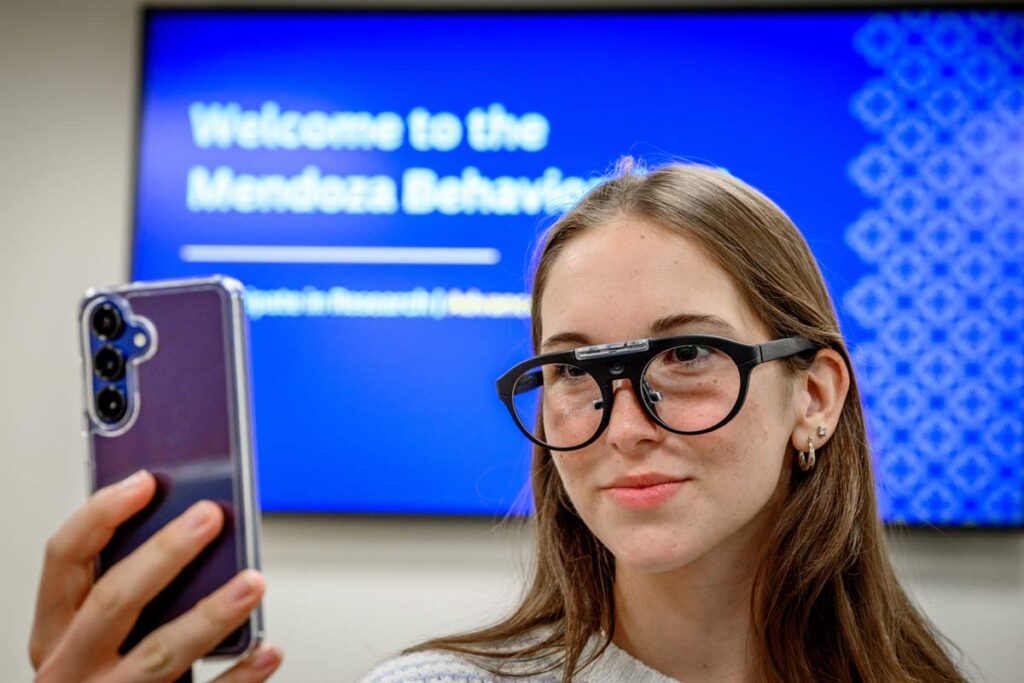 A student wears eye tracking glasses that link to a cell phone. The Behavioral Lab welcome slide is on a screen behind her.