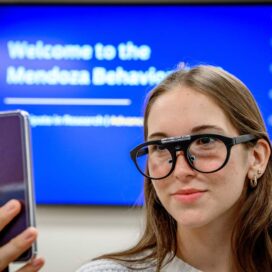 A student wears eye tracking glasses that link to a cell phone. The Behavioral Lab welcome slide is on a screen behind her.