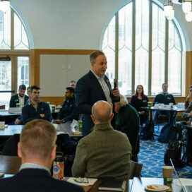 Nick Berente stands with a microphone in a classroom with audience members at tables around him.