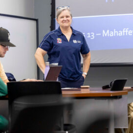 A smiling woman stands in front of students in the library classroom.